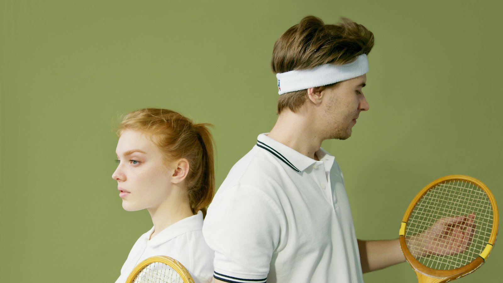 Caucasian couple in white tennis attire posing back to back with wooden rackets against a green backdrop.