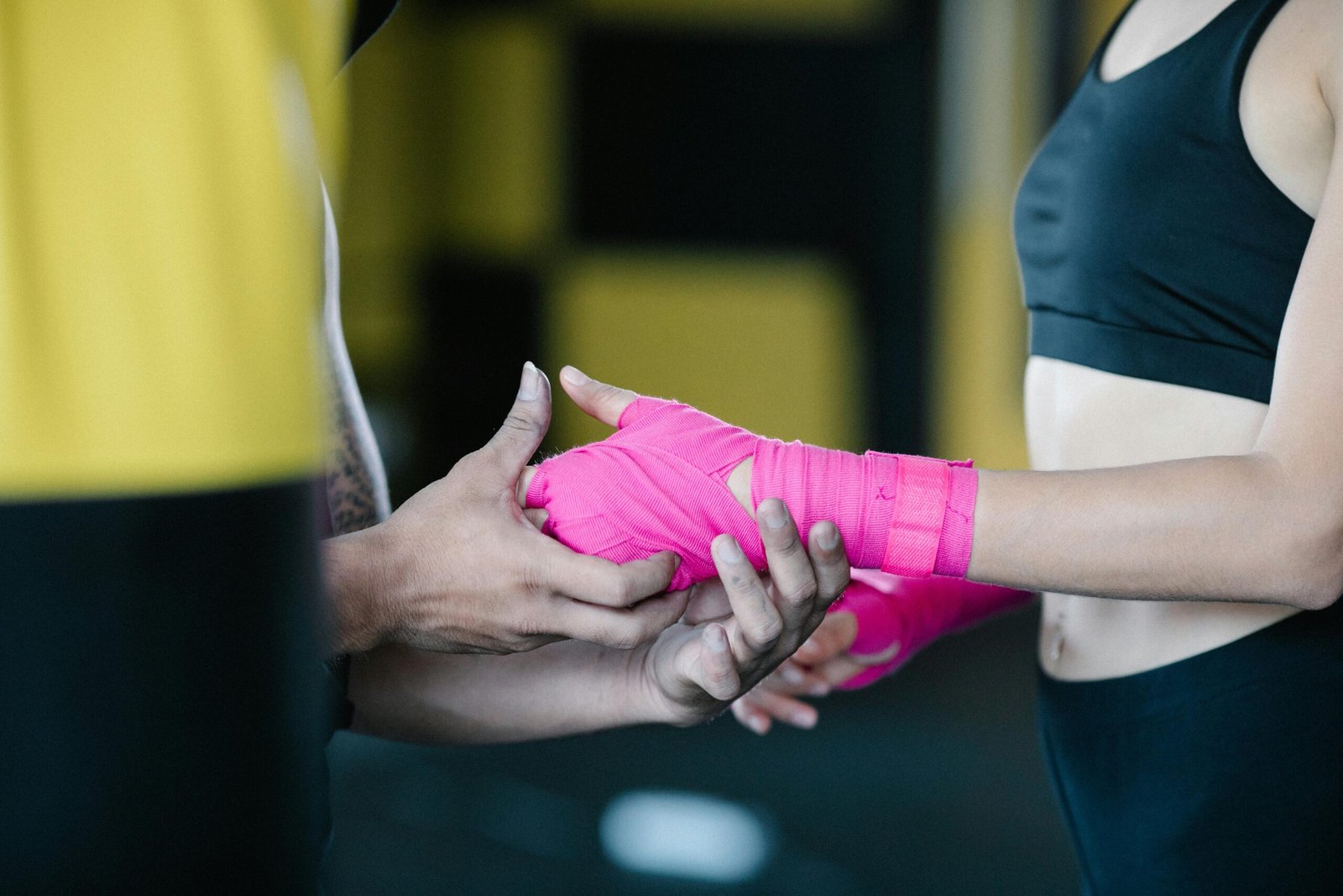 A woman receives assistance with pink hand wraps in an indoor gym setting, preparing for boxing practice.